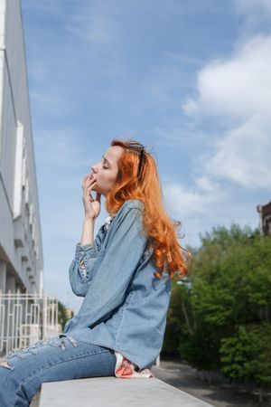 Side view of young woman with long red hair touching her mouth and sitting on balustrade near trees