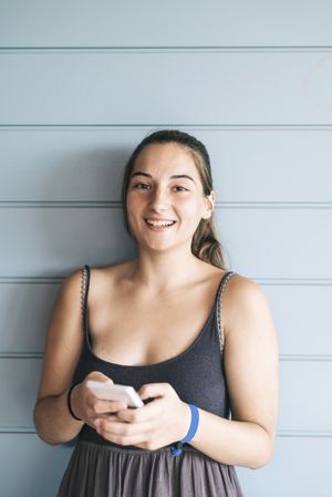 Happy teenage female looking up from cell phone while leaning against wood paneled wall