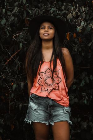 Woman in orange tank top and blue denim shorts standing near green leaves