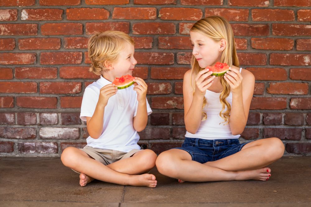 Cute Young Boy and Girl Eating Watermelon Against Brick Wall