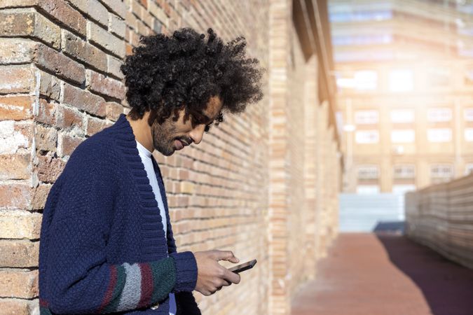 Curly haired man looking down at his smartphone while leaning on a brick wall outdoors on sunny day