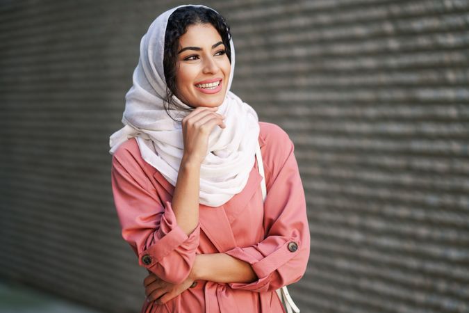 Happy female in headscarf and pink trench coat standing confidently with with one hand to her chin