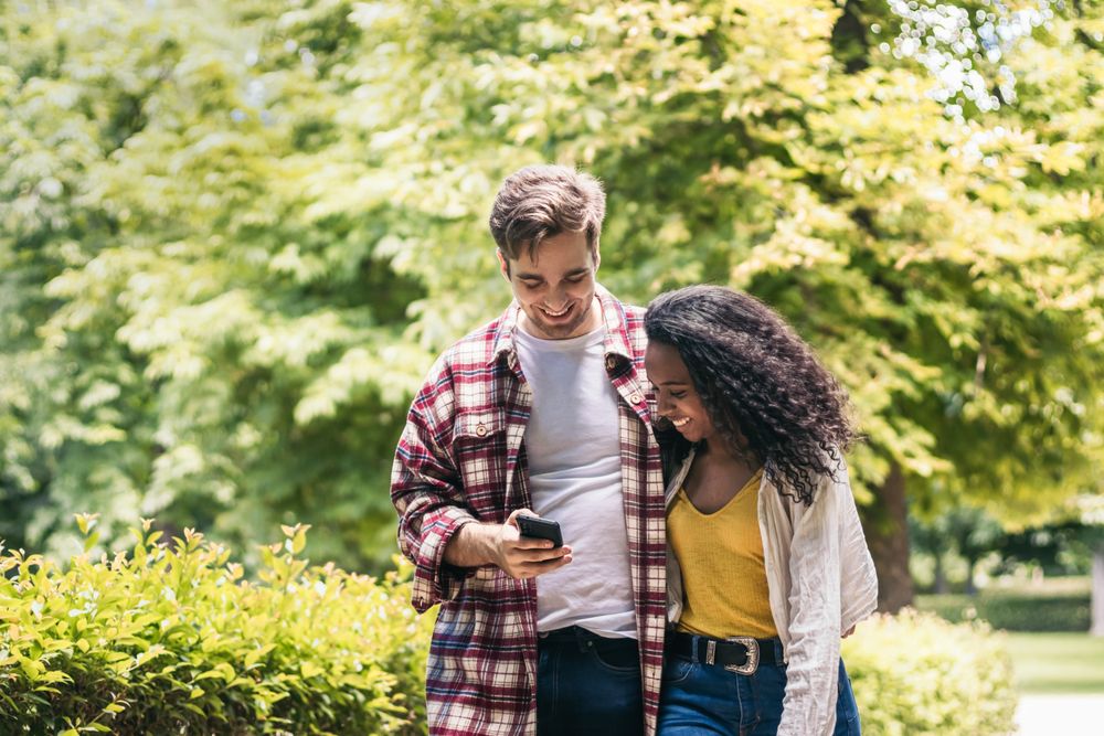 Happy multiracial couple taking a walk in the park using smartphone