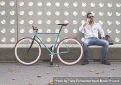 Male speaking on phone while sitting with bike parked in front of patterned cement wall 4OmNg0
