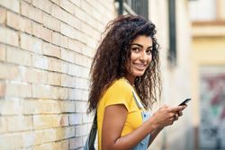 Arab woman looking back while texting with her smart phone outside in front of brick wall