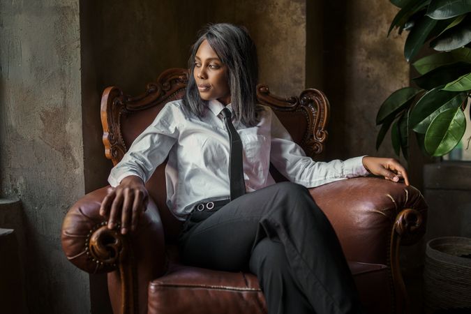 Portrait of a Black female businesswoman in trousers and tie sitting in a leather chair