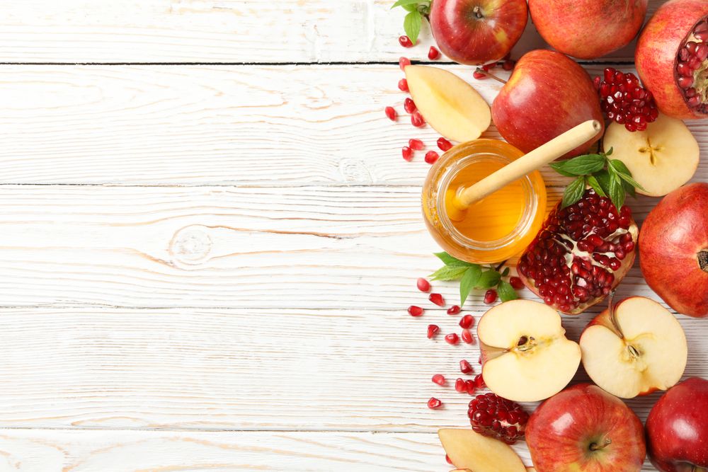 Top view of glass honey pot with dipper, pomegranate seeds, and an apple on right side of table