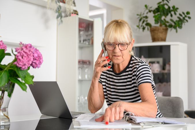 Worried old woman reading papers and having phone conversation at living room at home