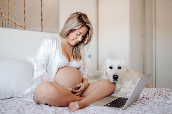 Cheerful pregnant woman using laptop and hugging dog