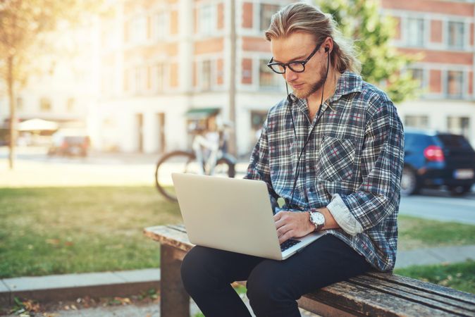 Man in glasses on his laptop on a bench in a city park