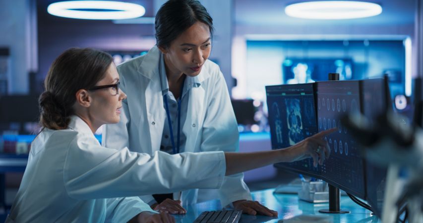 Asian And White Female Doctors Talking In Medical Research Center And Using Desktop Computer To Analyze MRI Scans Of Brain.