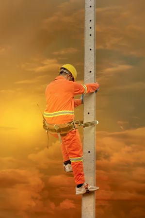Back view of man in orange overall and yellow bump cap climbing a pole