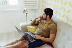 Young pensive male with laptop sitting on sofa
