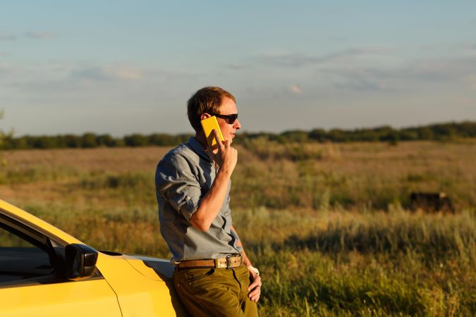 Male talking on his cell phone while leaning on front of stopped yellow car
