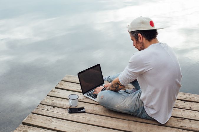 Man sitting on dock with laptop, smart phone and coffee