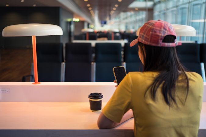 Asian woman browsing smartphone while drinking coffee at table under lighting lamp
