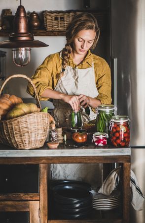 Young woman in rustic kitchen putting cucumbers in jar for pickling