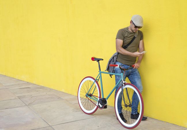 Male in hat and sunglasses standing next to yellow wall with bike and looking down at phone