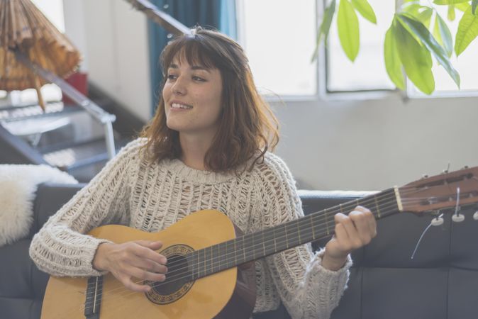 Content female strumming guitar in living room of bright loft with plants in background
