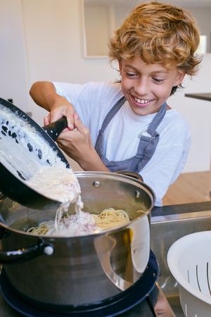 Smiling boy pouring carbonara sauce into spaghetti in kitchen