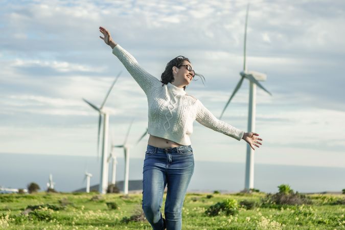 Excited woman in light sweater and denim pants standing beside wind turbines