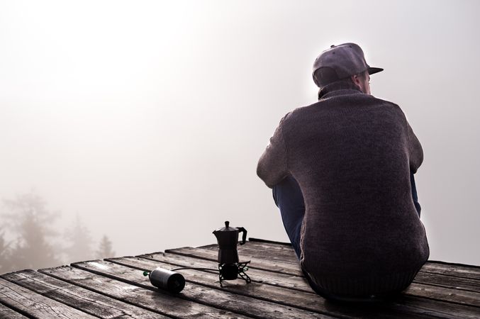Man in sweater sitting on wooden dock beside coffee pot