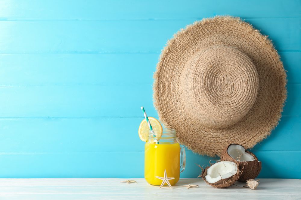 Straw hat, coconuts, fresh orange juice and starfishes on table against color wooden background, space for text. Summer vacation backdrop
