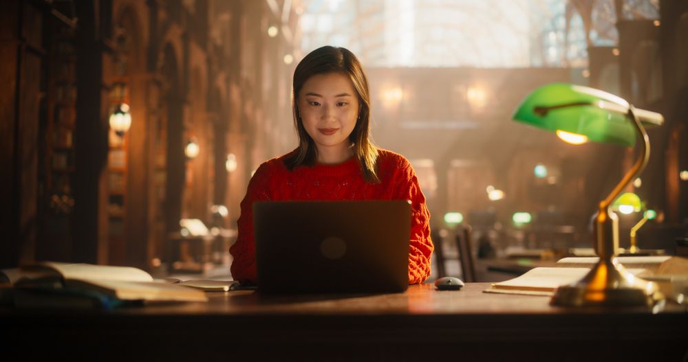 Dedicated Asian Female Student Engaged in E-Learning With Laptop in Classical Library.