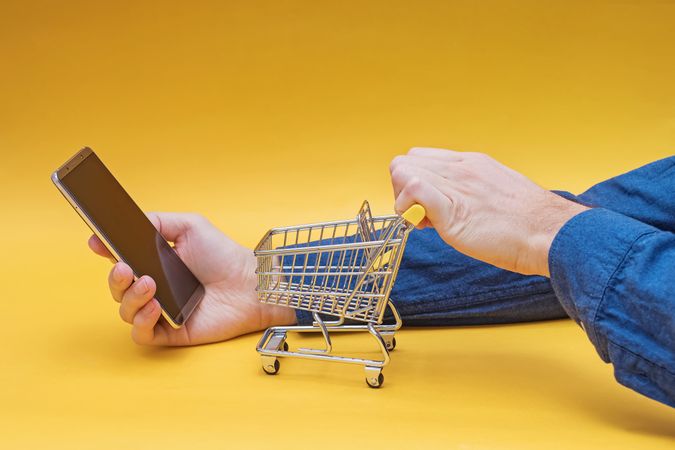 Hands Using Smartphone and Mini Shopping Cart on Yellow Background