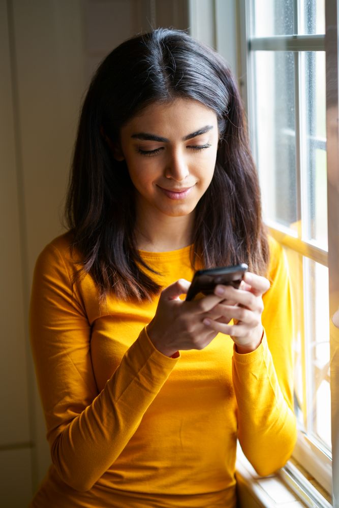 Female texting on phone next to window
