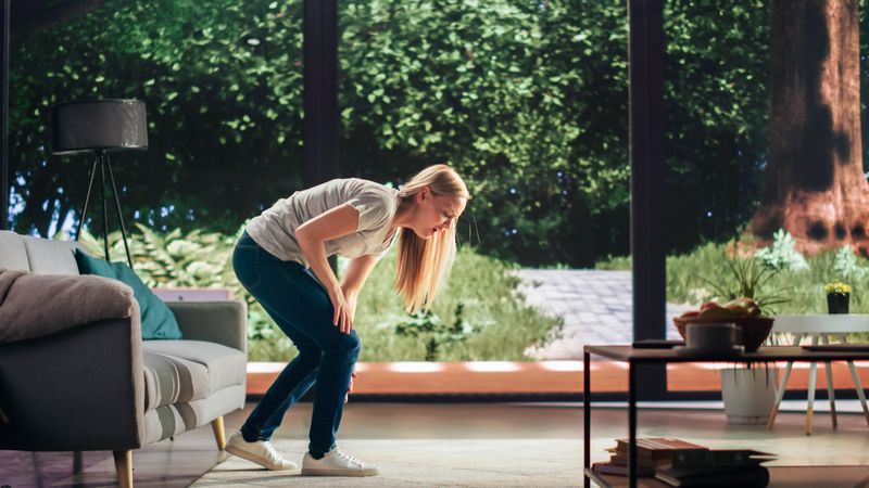 Young Adult Female Standing Up from a Couch at Home and Experiencing Discomfort in a Result of Leg Trauma or Arthritis. Joint and Knee Injury Concept.