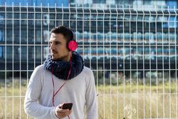 Young man walking in scarf leaning back on metallic fence and looking around holding smartphone