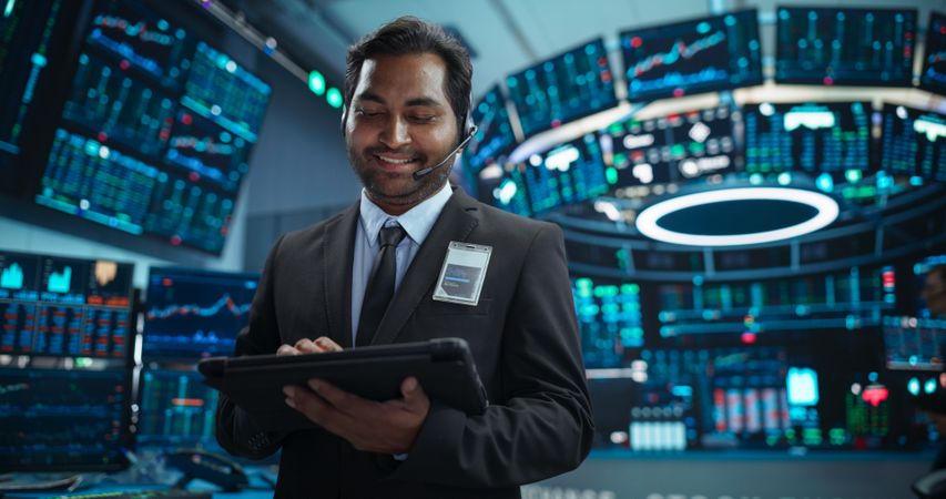 Portrait of a Young Indian Man Working in an International Stock Exchange Company: Happy Trader Using a Tablet Computer, Monitoring, Talking to a Corporate Business Partner in Headphones