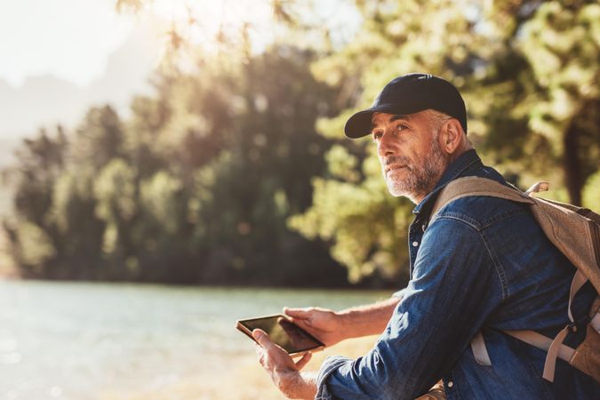Portrait of man sitting near a lake with backpack and digital tablet