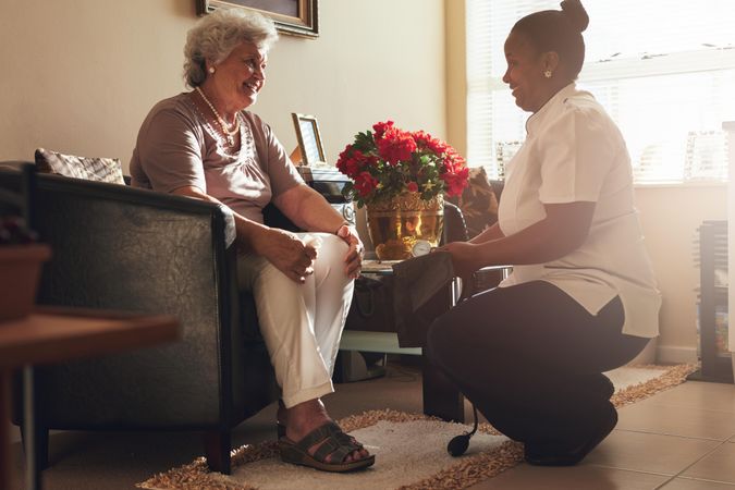 Mature woman sitting on a chair at home with female caregiver holding blood pressure gauge