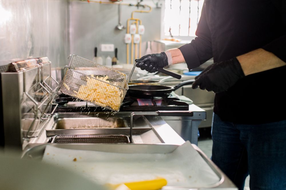 Crop anonymous male chef in gloves with deep fryer pan preparing French fries in kitchen of restaurant with utensils