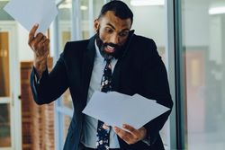 Overworked businessman in suit and tie with papers walking in office while speaking on smart phone