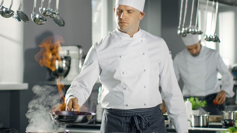 Professional Chef Fires up Oil on a Pan. Flambe Style Cooking. He Works in a Modern Kitchen with Lots of Ingredients Lying Around.