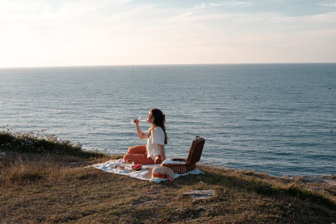 Woman with glass of drink on picnic mat looking at sea and mountains