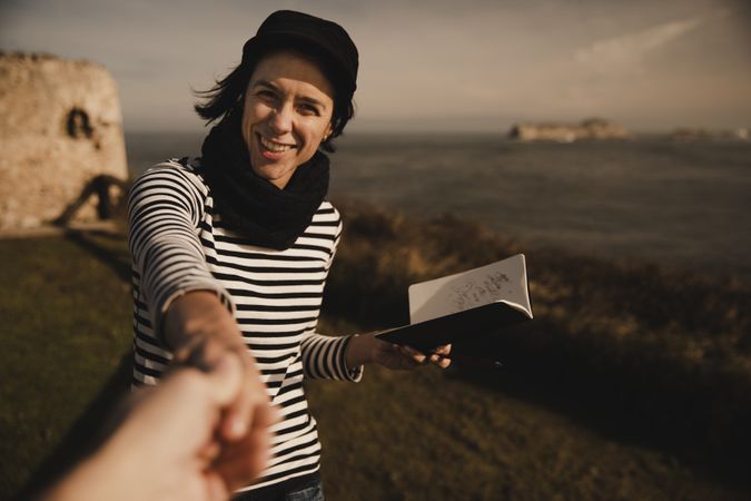Smiling woman with notebook holding hand on shore near water