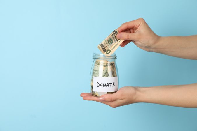 Female hand puts money in glass jar on blue  background