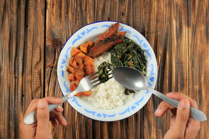 Top view of person with fork and spoon over Indonesian meal with meat, rice and vegetables