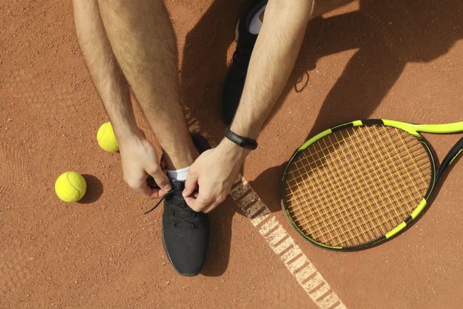 Man ties his shoelaces on clay court with racket and balls