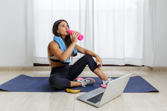 Young woman sitting on yoga mat beside computer drinking water