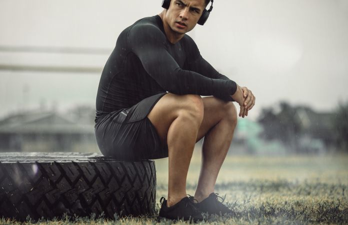 Healthy young man sitting on a big tire outdoors in the rain during workout