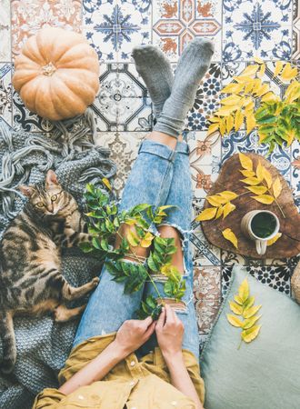 Woman with cat on blanket sitting on colorfully tiled balcony with fall leaves, squash, and mug