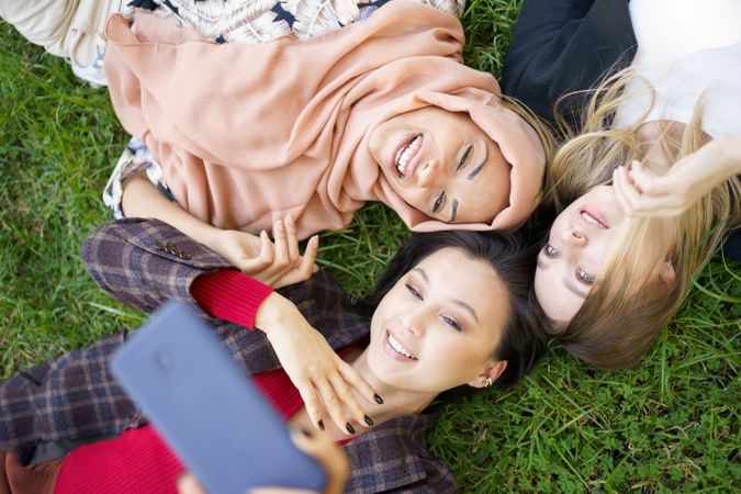 Looking down at three women lying on grass taking picture with smartphone