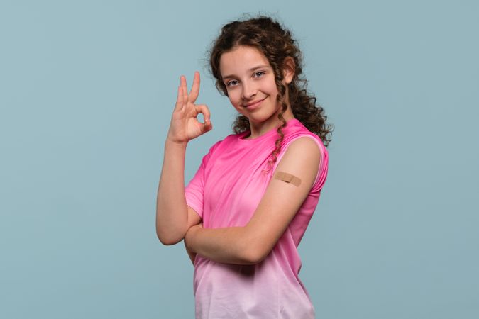 Smiling girl giving "OK" gesture, showing bandage on arm.