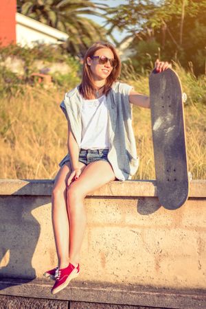 Young girl with skateboard sitting over the wall on summer