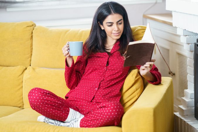 Woman sitting on sofa in red pajamas reading at home with book